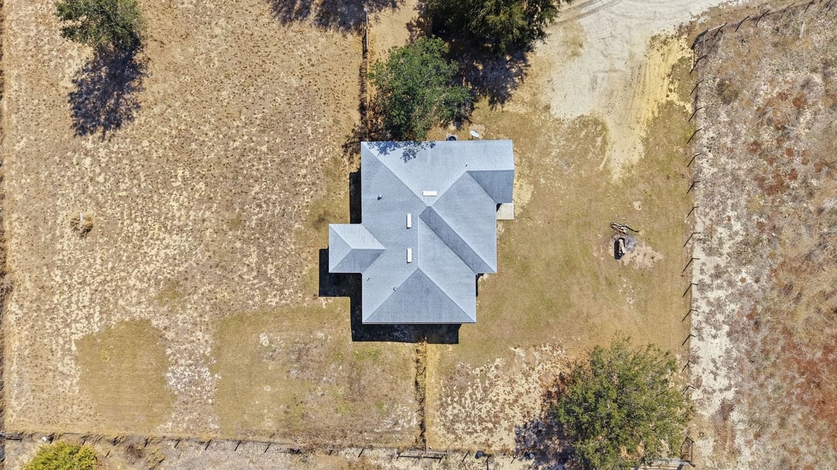 Aerial view of a residential house with gray roof surrounded by brown fields and scattered trees