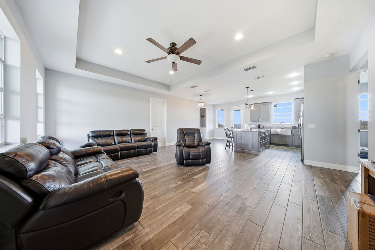 Spacious open-concept living room with brown leather furniture, wood plank flooring, ceiling fan, and large windows