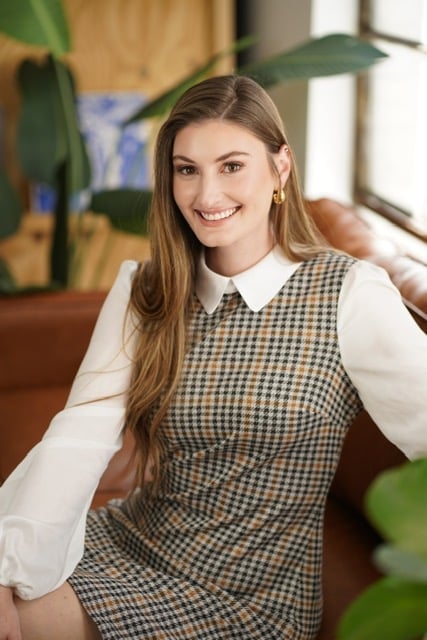 Smiling young woman with long brown hair wearing a checkered vest over a white long-sleeved shirt, seated near indoor plants